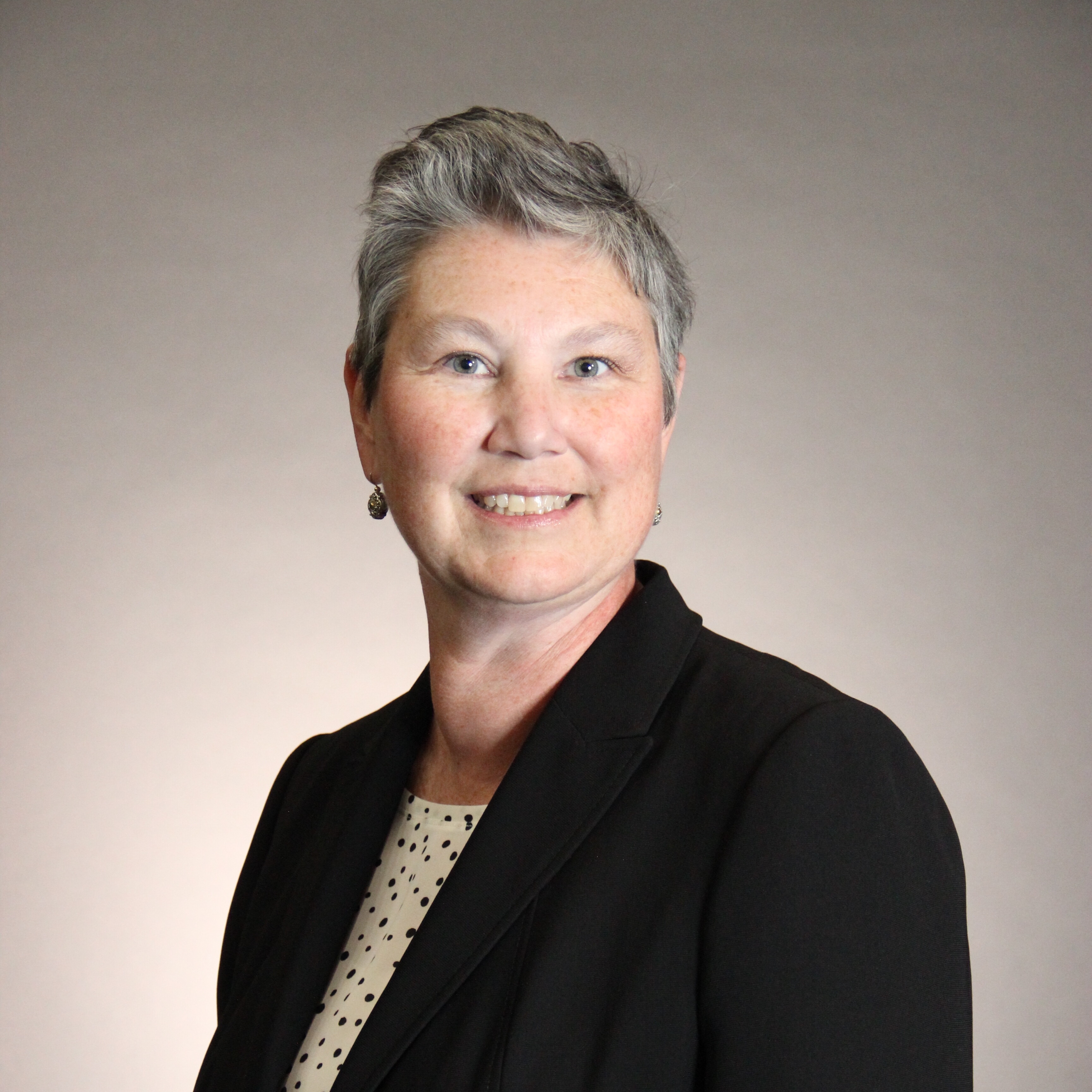 Carissa Uhrmacher white woman, middle aged, grey hair in a short styled cut, blue eyes and a nice smile wearing a black jacket and a white with black dots blouse, she is positioned in a traditional headshot, face to camera shoulders turned 3/4 to the left. She looks bright and engaged and positive