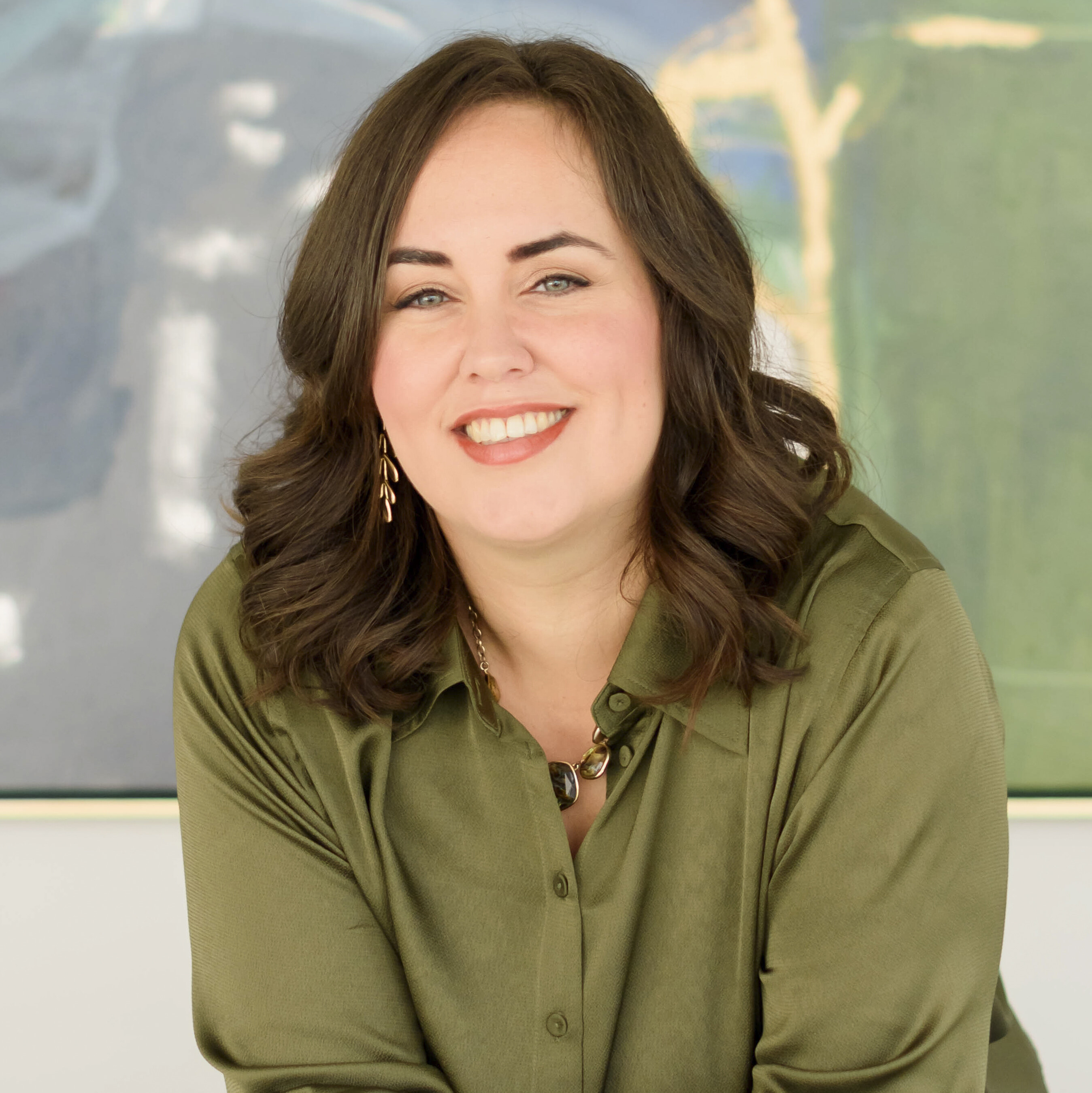 Trina Boos a woman with white skin, blue eyes and shoulder length brown hair, smiles warmly at the camera she is wearing an olive green buttoned and collared shirt and is posed in front of an abstract painting that has green, grey, blue, and sandy colors on it..