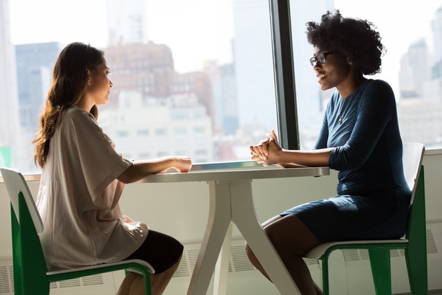 photo: two women sit at a table facing each other, talking and smiling over coffee